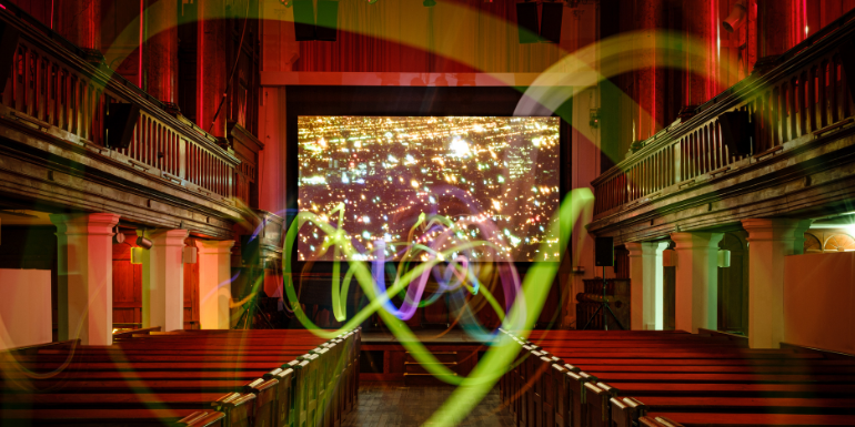 Image is of a cinema screen in a church. There is wooden pew seating on the bottom left and right with a centre aisle. There are wooden railings on the upper left and right. The screen has an image of a city at night - lots of colourful lights. The building is backlit with red lighting. There are artistic swirls of colour - green, blue, yellow, purple - overlaid on the image.