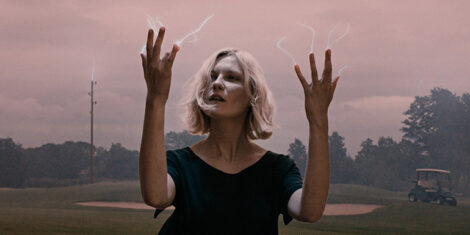 A white woman is holding her hands up and looking at her right fingertips. Electricity is arcing from all her fingertips. The sky is overcast, with a pink tinge. The woman is standing on a golf course. She is portrayed by Kirsten Dunst.