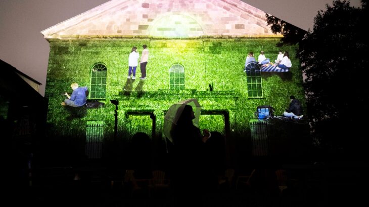 A projection of people on a field against the wall of a church. The silhouette of someone can be seen at the bottom of the image.