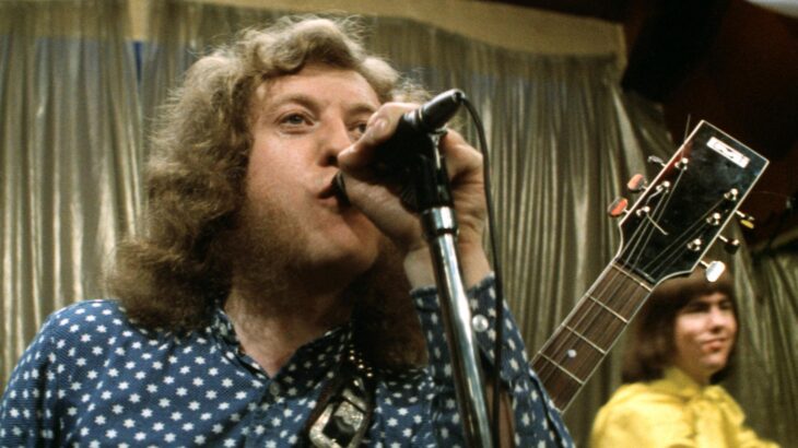 A man with frizzy hair and a guitar is singing into a microphone.